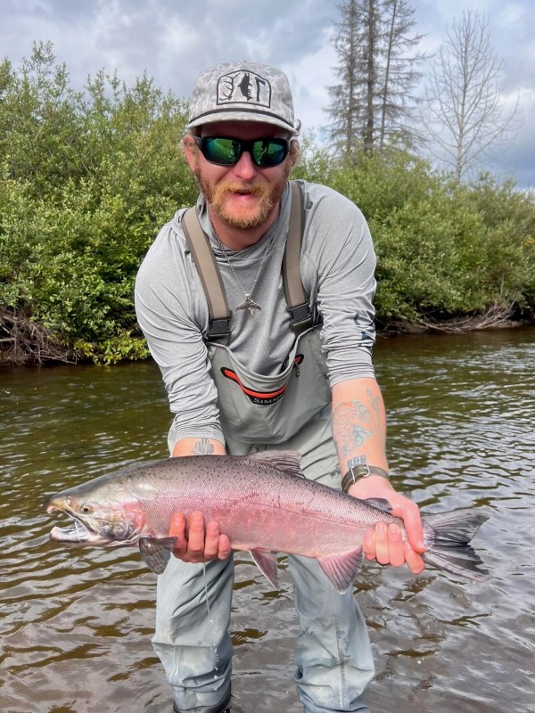 Man in waders holds a large fish in a river with greenery in the background.