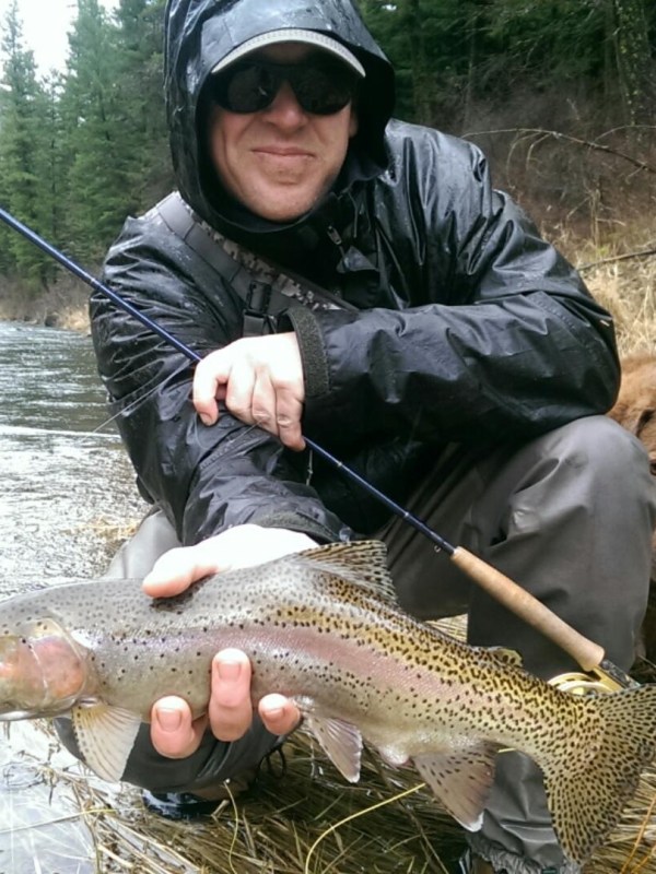 Person holding a large fish by a river, wearing rain gear, with a brown dog in the background.