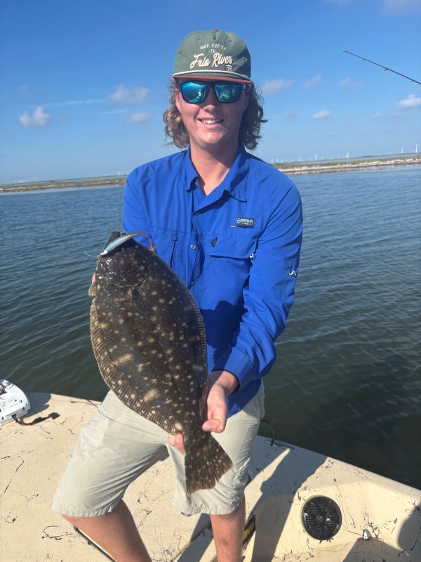 Person in blue shirt holding a flounder fish on a boat with water in the background.