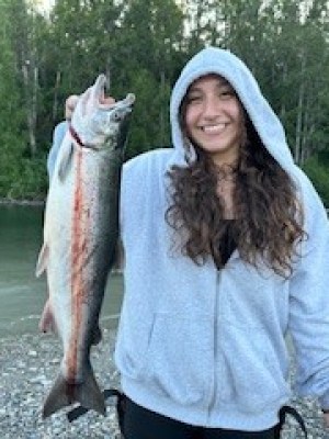 Person by a riverbank holding a large fish, wearing a gray hoodie and smiling.